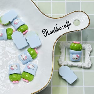 May include: A collection of miniature resin cactus figurines in blue pots with a cat face design. The cacti are green with pink flowers. The image also includes a white serving dish with gold trim and the word "Northcraft".