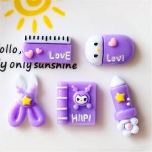 Può includere: Ciondoli in resina viola e bianca con vari design, tra cui un libro con la parola "Hiipi", un paio di forbici, un personaggio che dorme con la parola "Lovi" e un righello con la parola "Love".