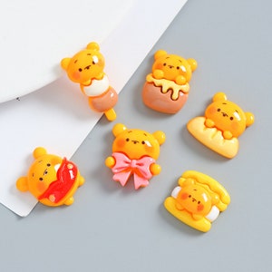 May include: Six yellow resin bear charms with different designs. The charms feature a bear holding a bow, a bear on a stick, a bear in a honey pot, a bear on a slice of bread, a bear in a hot dog bun, and a bear with a red heart.