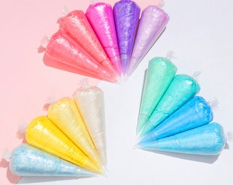 May include: A collection of colorful, pre-filled piping bags arranged in a fan shape. The bags contain various shades of frosting, including pink, purple, green, blue, and yellow. Each bag is sealed at the top, ready for use in baking and decorating.