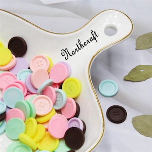 May include: Assortment of colourful plastic buttons in pastel shades of pink, yellow, blue, and green. The buttons are arranged in a white ceramic dish with a gold rim. The dish has the word "Northcraft" written on it in black cursive.
