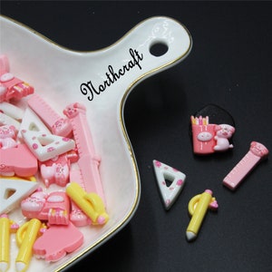 May include: Assortment of pink and white plastic school-themed buttons with pig designs. The buttons include rulers, pencils, protractors, and a pencil box with pencils. The buttons are in a white ceramic dish with a gold rim. The dish has the word "Northcraft" written on it.