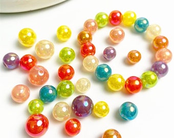 May include: A collection of iridescent glass beads in various sizes and colors, including yellow, red, green, and purple. The beads are scattered on a white surface, with some arranged in a circular pattern. The beads have a glossy finish.