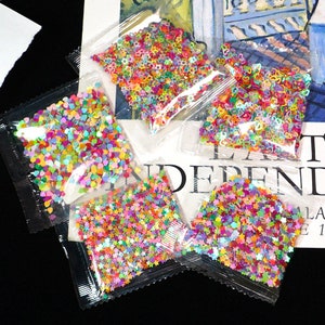 May include: Four small bags of colorful iridescent glitter. Each bag contains a different shape of glitter: stars, hearts, flowers, and a mix of shapes. The bags are clear plastic and are lying on a white surface with text.