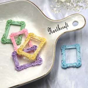 Peut inclure: Assortiment de cinq cadres photo décoratifs dans des tons pastel, y compris le vert, le rose, le jaune et le violet. Les cadres sont en plastique et présentent un design orné légèrement en relief. Les cadres sont présentés sur une assiette en céramique blanche avec un bord doré. L'assiette porte l'inscription "Northcraft" en écriture cursive noire.