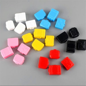 May include: Assortment of colourful miniature suitcase charms in various colours including pink, yellow, blue, red, black and white. The charms are made of plastic and have a suitcase design with a small heart on the front.