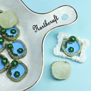 May include: A white serving tray with gold trim and the word "Northcraft" in black script. The tray holds several miniature pond-shaped decorations with blue water, green lily pads, and a stone border. A white framed pond decoration and a light green stone are also present.
