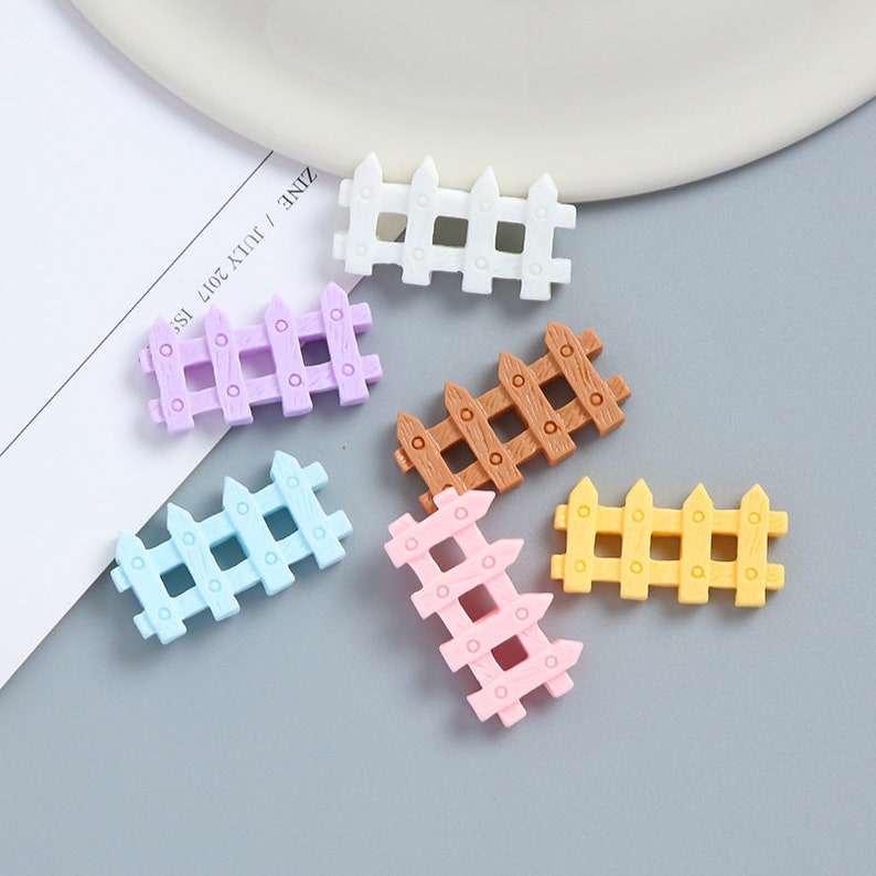 May include: Six miniature plastic fences in various colors including white, purple, brown, blue, pink, and yellow. The fences are all the same size and shape and have a simple design with vertical slats and horizontal rails.