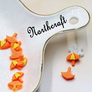 May include: A white ceramic dish with a gold rim, featuring the word "Northcraft" in black script. The dish contains several small orange and yellow umbrella-shaped charms. A separate white snowflake charm and two additional orange umbrella charms are on the white surface.