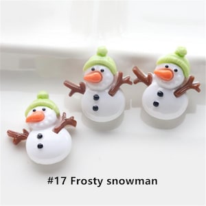 Pode incluir: Três pequenas figuras de boneco de neve brancas com chapéus verdes e braços de galho marrom. Os bonecos de neve têm narizes de cenoura laranja e olhos pretos. O texto #17 Frosty snowman está abaixo dos bonecos de neve.