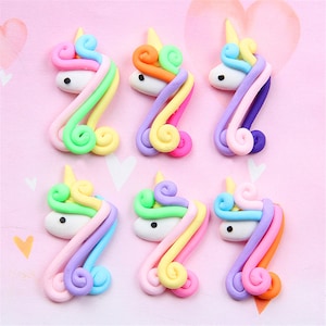Könnte beinhalten: Sechs bunte Einhornkopf-Anhänger aus Polymer Clay. Jeder Anhänger zeigt einen weißen Einhornkopf mit einer Regenbogenmähne und einem Horn.