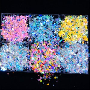 Op de afbeelding: Zes containers met iriserende glitter in verschillende kleuren en vormen, waaronder sterren, harten en bloemen. De glitter is gerangschikt in een rasterpatroon en bevindt zich in doorzichtige plastic containers.
