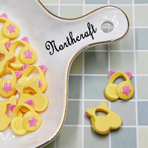 May include: A collection of yellow and pink resin charms shaped like a small purse with a star on the front. The charms are displayed on a white ceramic dish with the word "Northcraft" written on it.