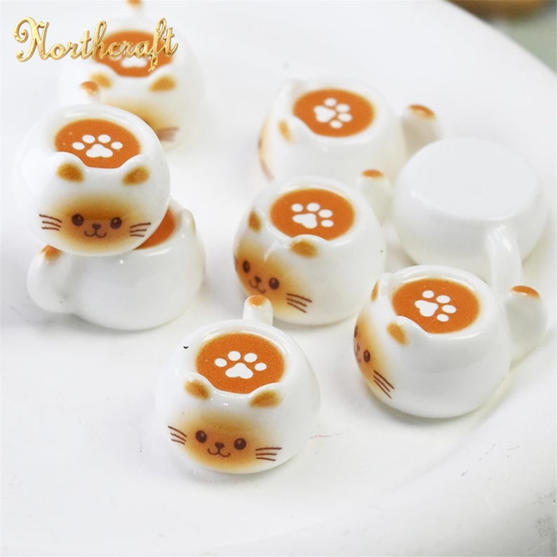 Puede incluir: Varias tazas de t&eacute; blancas de cer&aacute;mica con forma de gato y detalles en naranja. Cada taza presenta un dise&ntilde;o de huella de pata en el interior y una cara de gato en la parte delantera. El logotipo "Northcraft" es visible.