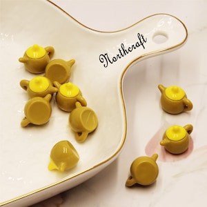 May include: A collection of nine miniature yellow teapots with yellow lids. The teapots are arranged on a white plate with a gold rim. The word "Northercraft" is written on the plate.