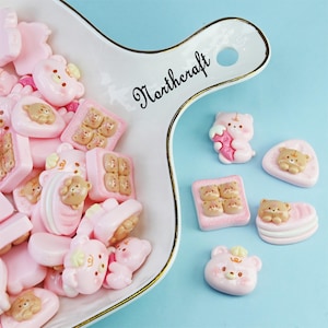 May include: A collection of pink resin bear-shaped charms and food-themed charms, including toast and macarons, are displayed on a white serving dish with gold trim. The dish is on a light blue surface. The word "Northcraft" is written on the dish.