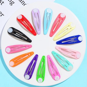 Peut inclure: Un ensemble de 15 pinces à cheveux colorées avec un bouton blanc à l'extrémité. Les pinces sont disposées en cercle sur une assiette blanche.