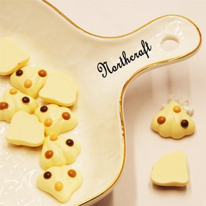 May include: A white ceramic dish with a gold rim, containing several small white and brown candy-like decorations. The dish has the word "Northcraft" written on it in black.
