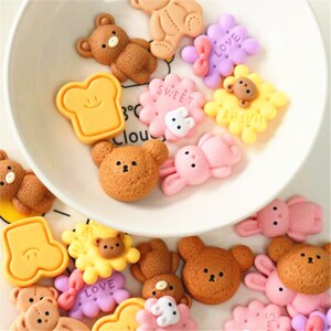 May include: Assortment of colorful resin charms featuring cute cartoon bears, bunnies, and bread slices. The charms have various designs and colors, including pink, yellow, brown, and purple. The charms are perfect for adding a touch of whimsy to crafts, jewelry, and other projects.