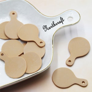 May include: A group of 10 beige, round, flat, wooden, craft embellishments with a small hole at the top. The embellishments are in a white ceramic dish with a gold rim. The dish has a handle and the word "Northcraft" is written on the handle.