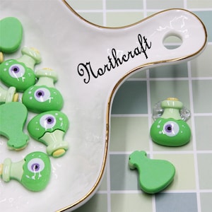 May include: Green and white plastic potion bottle charms with a single purple eye on each bottle. The charms are on a white plate with a gold rim. The plate has the word "Northcraft" written on it in black cursive.