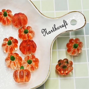 May include: A collection of orange and clear glass pumpkin decorations with green stems, displayed on a white serving dish with gold trim. The dish has the word "Northcraft" on it. These pumpkins are a fall or autumn theme.