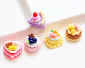 May include: Six miniature cake charms in various colors and designs, including a purple cake with a cherry on top, a yellow cake with orange slices, a pink cake with a chocolate bar and blueberries, a pink heart-shaped cake with a chocolate bar and yellow sprinkles, a yellow cake with pink and white frosting and a brown cake with yellow sprinkles.