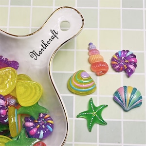 May include: A white serving dish with gold trim holds colourful resin sea-themed embellishments. The dish has the word "Northcraft" in script. Scattered around are a green starfish, a striped seashell, and other decorative shells.