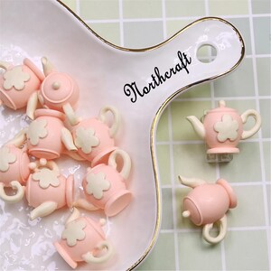 Può includere: Una collezione di piccoli ciondoli a forma di teiera rosa con dettagli di fiori e manici bianchi. I ciondoli sono disposti su un piatto da portata bianco con bordo dorato. La parola "Northcraft" è scritta in nero sul piatto.