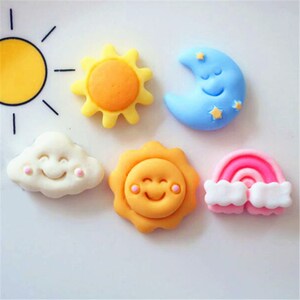 May include: Five colourful, smiling, cartoon-style decorations, including a sun, a cloud, a moon, a rainbow, and a sun with a face. The decorations are made of a soft, pliable material, possibly fondant or gum paste.