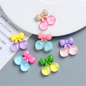 May include: Six pairs of colorful resin cherry charms with bows. Each charm features two round cherries with a bow on top. The colors include pink, purple, blue, yellow, green, and beige.