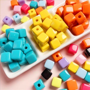 Könnte beinhalten: Sortiment an bunten Plastikwürfelperlen in verschiedenen Farben, darunter Gelb, Orange, Blau, Rosa und Lila. Die Perlen sind auf einem weißen Teller mit weißem Hintergrund angeordnet.