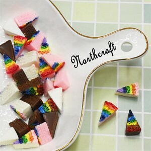 Puede incluir: Un plato blanco con borde dorado contiene mini rebanadas de pastel en varios colores, incluyendo arcoíris, chocolate y rosa. El plato está sobre una superficie a cuadros verde claro y blanco. La palabra "Northcraft" está escrita en cursiva negra.