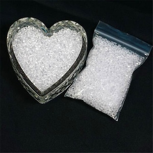Puede incluir: Dos recipientes llenos de cuentas redondas transparentes y translúcidas. Un recipiente es un plato de vidrio en forma de corazón, y el otro es una bolsa de plástico transparente.