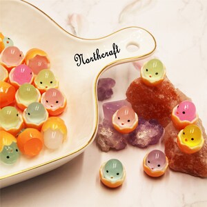 Peut inclure: Une collection de petits objets ronds et colorés, chacun ressemblant à un petit fruit avec un visage. Les perles sont de différentes couleurs, dont orange, rose, vert et transparent, disposées sur un plat blanc et dispersées parmi des cristaux.