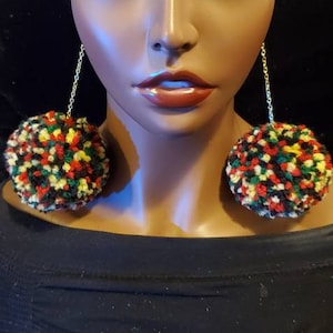 May include: A pair of dangle earrings with large, colorful pom-poms. The pom-poms are made of yarn in shades of red, yellow, green, and black.