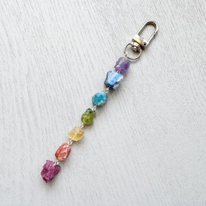 Könnte beinhalten: Ein Schlüsselanhänger mit einer Reihe von farbigen, rohen Edelsteinperlen in einem Regenbogenspektrum. Die Edelsteine sind mit einer silberfarbenen Kette aufgefädelt und an einem Metallverschluss befestigt. Die Edelsteine umfassen Lila, Blau, Grün, Gelb, Orange und Rosa.