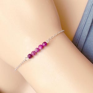 May include: A delicate silver chain bracelet with a row of small, round, purple beads. The beads are graduated in size, with the largest bead at the center of the bracelet.