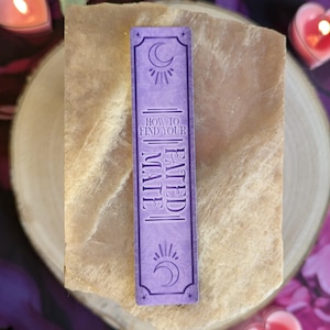 Può includere: Un segnalibro lavanda con la scritta "HOW TO FIND YOUR FATED MATE". Il segnalibro presenta un disegno di sole e luna crescente in alto e in basso. Lo sfondo include una fetta di legno e candele accese.