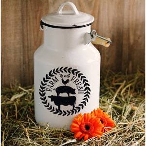 Pu&ograve; includere: Una latta di latte smaltata bianca con un design in bianco e nero che dice "Farm Fresh" con un gallo, un maiale e una mucca. La latta ha un manico di legno e si trova su un letto di fieno.