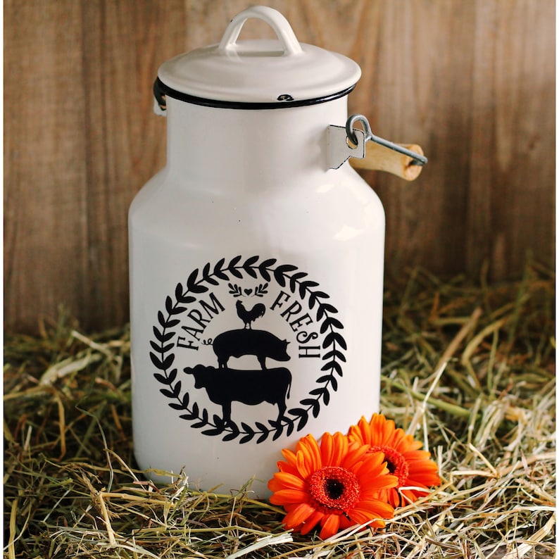 Pu&ograve; includere: Una latta di latte smaltata bianca con un design in bianco e nero che dice "Farm Fresh" con un gallo, un maiale e una mucca. La latta ha un manico di legno e si trova su un letto di fieno.