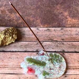 May include: White resin incense holder shaped like a hamsa hand with green and red floral accents and iridescent glitter. A brown incense stick is burning in the holder.