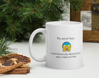 Puede incluir: Taza de café de cerámica blanca con una cara de dibujos animados negra y amarilla y el texto "Mi peor miedo... es desarrollar espontáneamente un nuevo miedo irracional."