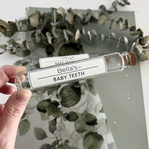 May include: A clear glass vial with a cork stopper and a white label that reads "Bella's Baby Teeth". The vial is on a green and white background with a floral pattern.