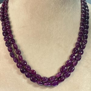 Könnte beinhalten: Eine dreireihige Halskette aus polierten, oval geformten, tiefvioletten Edelsteinen. Die Halskette wird auf einer neutralfarbenen Schaufensterpuppe präsentiert, die die satte Farbe und Textur der Steine hervorhebt. Die Edelsteine sind aneinandergereiht und erzeugen einen mehrschichtigen Effekt.