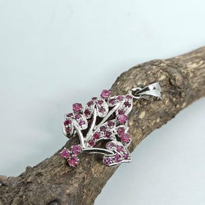 May include: A silver pendant shaped like a tree branch, set with small, round, pink gemstones. The pendant is displayed on a piece of weathered wood, set against a pale blue backdrop.