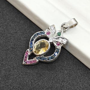 May include: A silver pendant with a leaf-like design. The pendant features a large, oval, yellow gemstone at the centre, surrounded by smaller, colourful gemstones in shades of blue, pink, green, and white. The pendant has a loop for a chain.