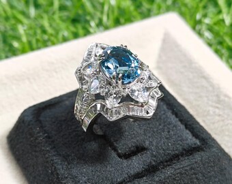 May include: A silver ring with a large, light blue gemstone at the center, surrounded by smaller clear stones. The ring has an intricate, floral-like design with multiple layers of stones. The ring is set on a black velvet surface against a blurred green background.