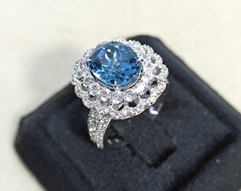 May include: A silver ring featuring a large, oval, light blue gemstone surrounded by a halo of smaller, clear gemstones. The ring's band is also adorned with small, clear gemstones. The ring is displayed on a black surface.