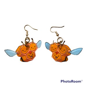 May include: A pair of earrings featuring a cartoon bee design. The bee is made of orange and pink paper and has a light blue wing on each side. The earrings are attached to silver hooks.